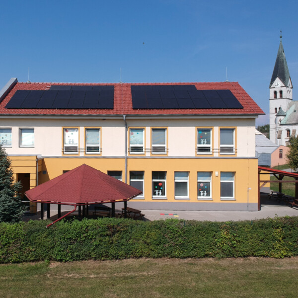 Two-story beige-yellow building with large dark solar panels on the red-tiled roof and a red gazebo in front; church spire visible in the background.