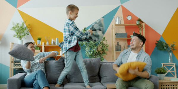 Child in a plaid shirt leaps with a pillow toward his smiling mom on a gray sofa, while dad sits with a yellow cushion in a bright, geometric living room.