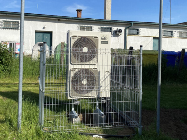 Outdoor HVAC unit with two large fans behind a chain-link fence in a grassy area by a building 0