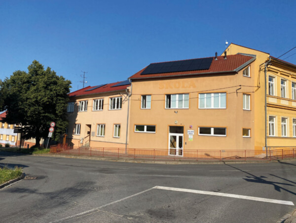 Two-story peach-colored school building with solar panels on the roof, beside a quiet street under a clear blue sky.