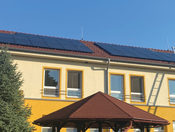 Two-story building with solar panels on the roof, yellow window frames, and a brown gazebo in front under a blue sky.