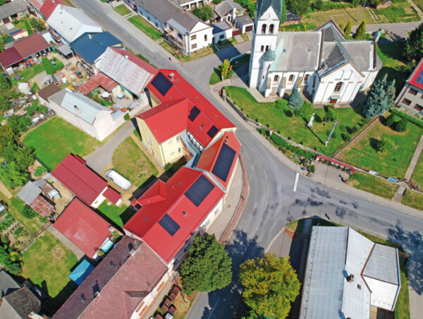 Aerial view of a small town with red-roofed houses, a white church, and winding streets beneath greenery.